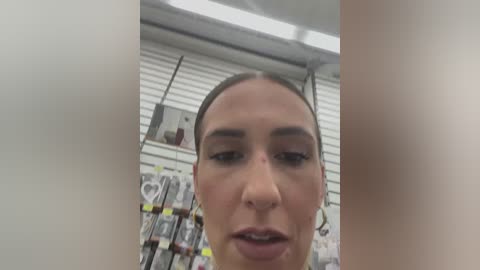 Media: Video of a woman with light skin and dark hair in a ponytail, wearing a yellow shirt, standing in a store with white shelves and products in the background.