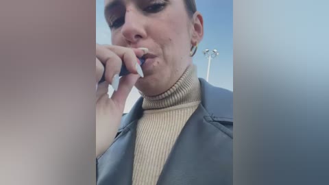 Media: A video of a woman with fair skin, wearing a beige turtleneck and a grey jacket, biting her fingernail. Background shows a clear blue sky and a football stadium.