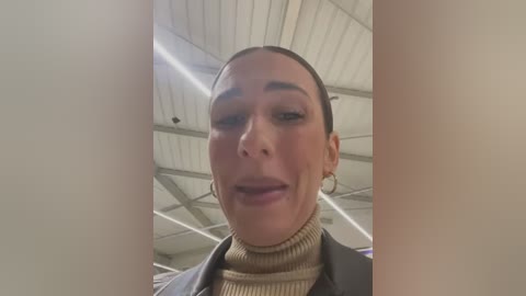 Media: Video of a woman with a light skin tone and short, slicked-back hair, wearing a beige turtleneck sweater and a black blazer. The background shows a modern, industrial ceiling with white panels and exposed beams.