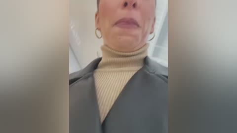 Media: A video of a woman's upper body in a gray coat with a beige turtleneck sweater. Her face is partially visible, wearing hoop earrings. The background is blurry, likely an indoor setting.