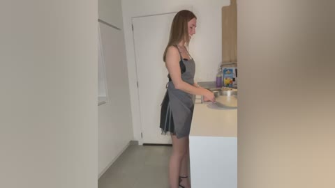 Media: A video of a slender, light-skinned woman with long brown hair in a gray dress, standing in a minimalist kitchen with white walls and wooden cabinets.