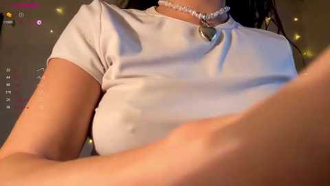 A video of a woman with fair skin wearing a tight, white crop top, a heart pendant necklace, and a choker, seated indoors with a dimly lit, warm-toned background.