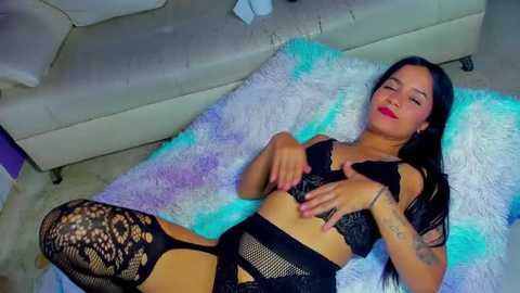 Media: Video of a Latina woman with long black hair, light skin, and medium breasts, lying on a white rug, wearing black lace lingerie and fishnet stockings. She has tattoos on her arms.