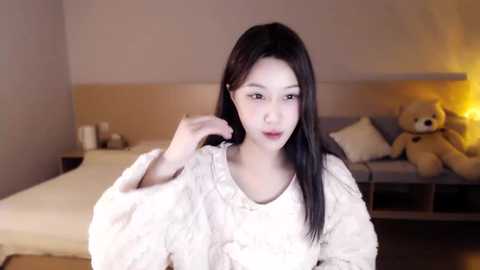 Media: A video of an East Asian woman with straight black hair, fair skin, and a slender physique, wearing a white fluffy robe, sitting on a bed with teddy bears in a softly lit, minimalist bedroom.