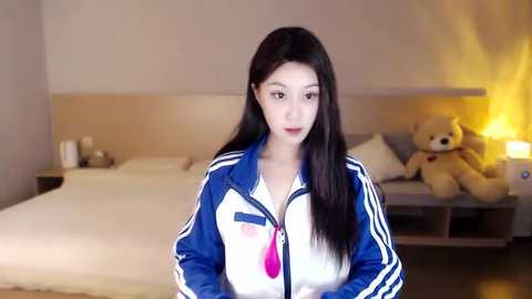 Media: Video of an Asian woman with long black hair, wearing a blue Adidas track jacket, standing in a modern bedroom with a bed, teddy bears, and a warm lamp.