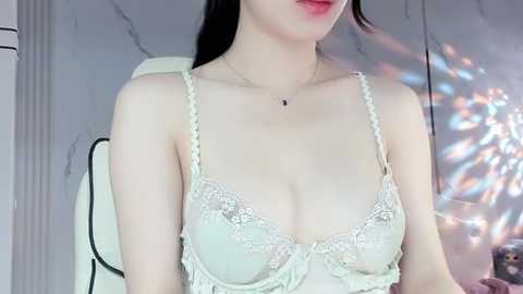 Media: Video of a slender, fair-skinned Asian woman with straight black hair, wearing a delicate, lacy white bra with floral patterns. She has a small, delicate necklace. Background includes a marble wall and a blurred, colorful light display.