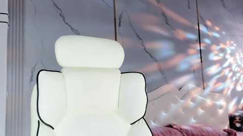 Media: Video of a modern white leather recliner chair with a tufted headrest, set against a marble-patterned wall with soft, pink and blue light reflections.