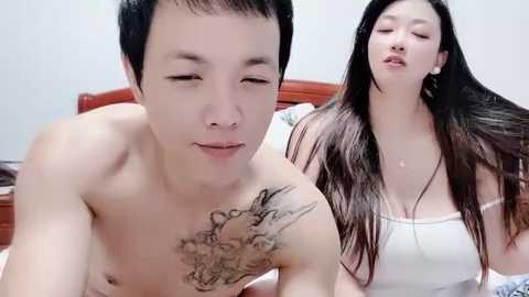 Media: Video of an Asian man and woman in bed, both topless. Man has a tattoo on his chest, woman wears a white tank top, both appear relaxed. Background shows a wooden headboard.