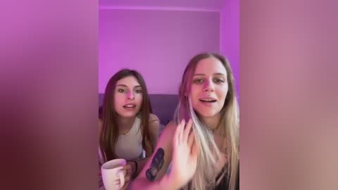 Media: Video of two young women, one with long brown hair and a white top, holding a cup, and the other with long blonde hair, waving, in a dimly lit room with purple lighting.