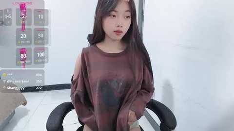 Media: Video of an Asian woman with long black hair, wearing a loose, dark purple sweater, sitting on a black chair in a modern, minimalist room.