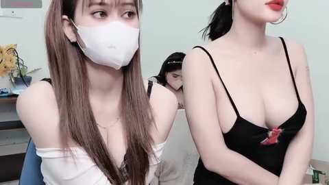 Media: Video of two Asian women in a room; one with long brown hair in a white mask, white top, and blue pants, and the other with black hair, black lingerie, and a red bow.