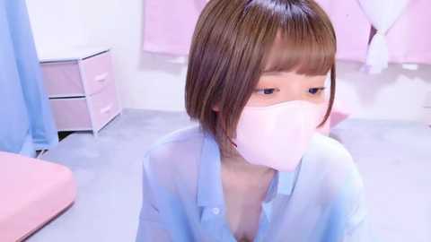 Media: Video of a young Asian woman with straight, shoulder-length brown hair, wearing a light blue shirt and a pink surgical mask, sitting on a light gray floor in a sterile, pink and white room.