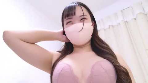 Media: A video of an East Asian woman with long black hair, wearing a pink lace bra, and a white face mask, posing in a bright, minimalist room with white curtains.