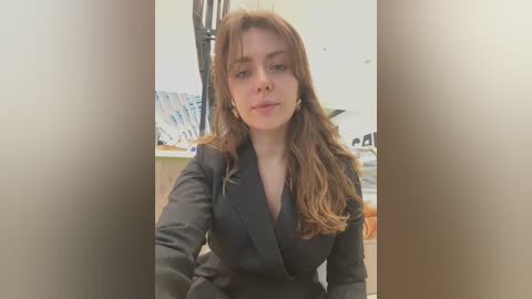 Media: Video of a Caucasian woman with long brown hair, wearing a black blazer, standing indoors with a blurred background featuring a ceiling fan and a table.