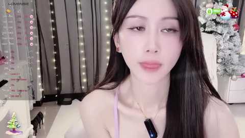 Media: Video of an Asian woman with long black hair, fair skin, and a slender figure, wearing a light pink top and a black necklace. Background includes a decorated Christmas tree and a white table with a small, colorful ornament.