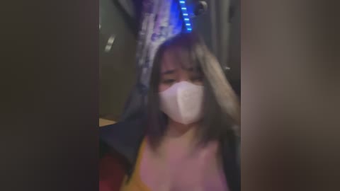 Media: Video of an Asian woman with shoulder-length black hair, wearing a yellow tank top and black jacket, and a white face mask, sitting on a subway train with dim lighting and blue LED lights.