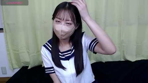 Media: A video of a young East Asian woman with straight black hair and fair skin, wearing a white sailor-style shirt, black mask, and sitting on a black couch against a pale green curtain.