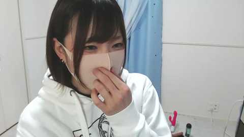 Media: Video of an Asian woman with short black hair, wearing a white hoodie and white face mask, covering her mouth with one hand, in a sterile medical setting with blue curtains and white walls.