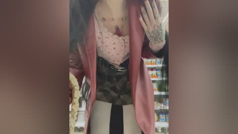 Media: Video of a person in a grocery store, wearing a pink lace bra, black jacket, and camo pants. Their hands are visible with tattoos. Background shows shelves filled with products.