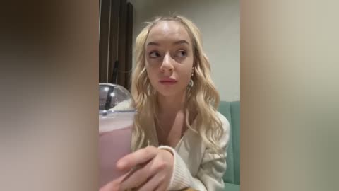 Media: A video of a young, fair-skinned blonde woman in a beige sweater, holding a pink smoothie with a straw, indoors near a green sofa.