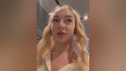 Media: Video of a young Caucasian woman with wavy blonde hair, fair skin, and pink lips, wearing a light-colored shirt, indoors with a modern ceiling and lighting.