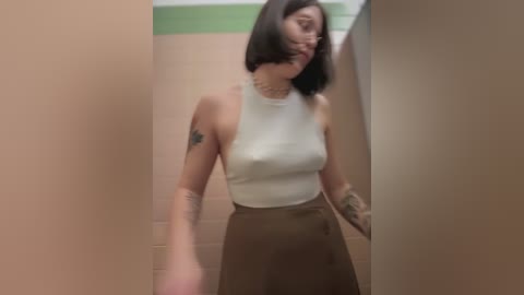 Media: A video of a woman with short black hair, medium skin tone, wearing a white tank top and brown skirt, standing in a beige-tiled bathroom with a green trim.