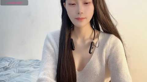 Media: Video of a young East Asian woman with long, straight black hair, wearing a light grey, knitted V-neck sweater and large, dark sunglasses. She has a calm expression. Background includes a light-colored wall and a messy bed with blue sheets.