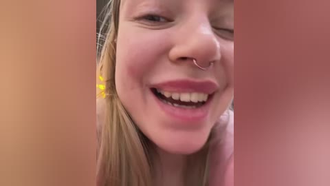 Media: Video of a young woman with fair skin, smiling with a visible septum piercing, wearing a yellow flower in her hair. The background is blurred and out of focus.