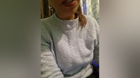 Media: A video of a woman with light skin, wearing a light gray knitted sweater, her mouth slightly open, and a light-colored background with a blurry pattern.