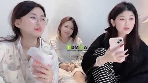 Media: Video of three East Asian women with light skin, straight black hair, and glasses, wearing pajamas, holding facial masks, smiling in a cozy, well-lit room.