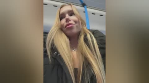 Media: A video of a smiling, blonde woman in a dark jacket, taken from a low angle, with a blurred, blue bus interior background.