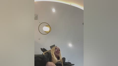 Media: Video of a young woman with long blonde hair, smiling, wearing a black graduation gown, standing in a modern, white-walled room with a circular ceiling light and recessed lighting.