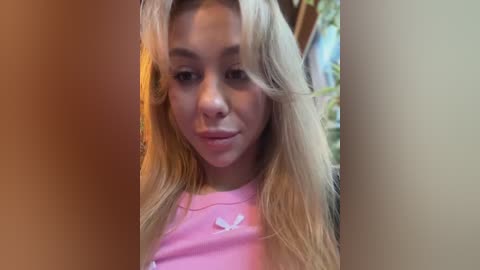 Media: A video of a young, light-skinned woman with long, wavy blonde hair, wearing a pink t-shirt with a white flower graphic, looking slightly to the side. The background is blurred and out of focus.