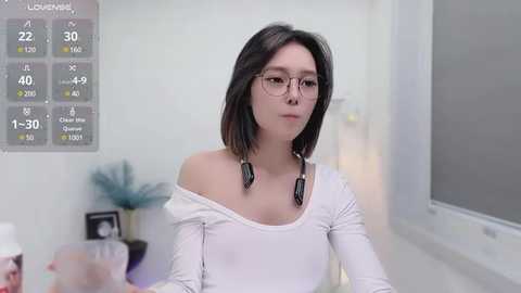 Media: A video of a young woman with straight, dark hair and glasses, wearing a white off-shoulder top, seated in a modern office. A wall-mounted thermometer is visible in the background.