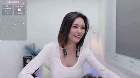 Media: Video of a young woman with straight, shoulder-length black hair, wearing a white long-sleeve top and black geometric necklace, seated in a modern, minimalist room with white walls and a large window.