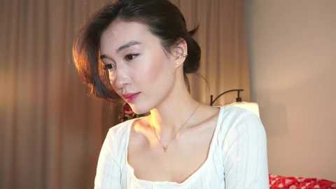 Media: Video of an Asian woman with fair skin, dark hair, and red lipstick, wearing a white lace top, standing indoors against beige curtains.