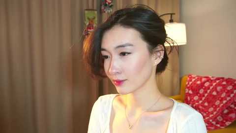 Video of a young East Asian woman with fair skin, dark hair, and light makeup, wearing a white cardigan, in a softly lit room with beige curtains, a red floral pillow, and a table lamp.