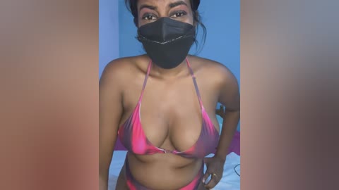 Media: Video of a South Asian woman with medium brown skin, wearing a pink and black bikini, black face mask, and black hair in a bun, indoors.