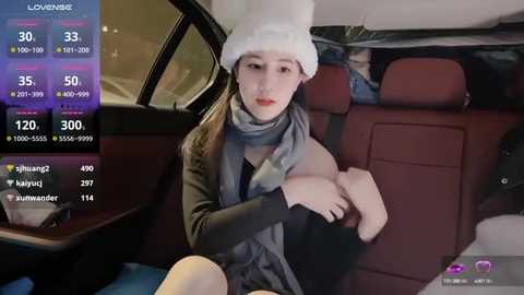 Media: Video of a young Asian woman in a car, wearing a white beanie, gray scarf, and black top, revealing cleavage. Background shows car interior with passengers, and social media app interface.