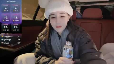 Media: Video of a young Asian woman with fair skin, wearing a white knitted hat and dark jacket, drinking water inside a car, with a digital weather app on the left screen.