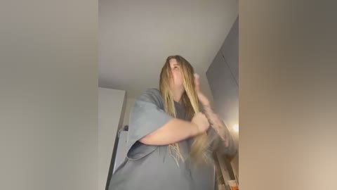 Media: Video of a young woman with long, blonde dreadlocks, wearing a loose, grey t-shirt, standing in a dimly lit room with white walls and a door visible in the background.