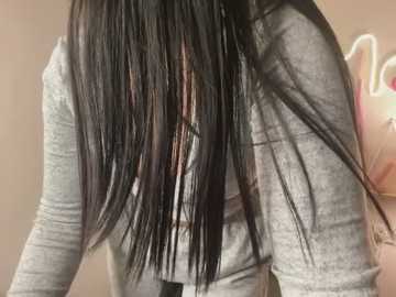 A close-up video of a person with long, straight, black hair styled with thin, light-colored streaks. They are wearing a gray, long-sleeved top, and the background is a blurred, light beige wall.