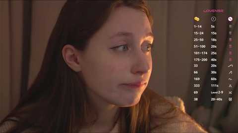 Video of a young Caucasian woman with fair skin, straight brown hair, and a neutral expression. She wears a beige sweater. Background is blurred, suggesting an indoor setting.