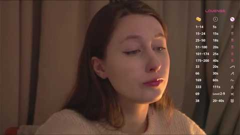 Media: Video of a young woman with light skin and long brown hair, wearing a beige sweater, in a dimly lit room with beige curtains. Her eyes are half-closed, and she has a serene expression.