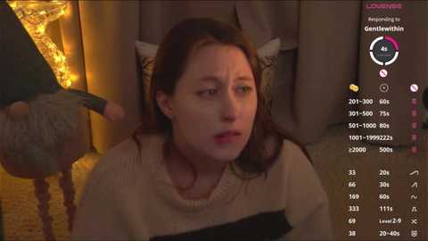 Video of a young, fair-skinned woman with long brown hair, wearing a cream sweater, sitting in a dimly lit room with warm, yellow string lights. She appears anxious, with her mouth slightly open.