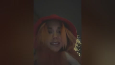 Media: A video of a woman with shoulder-length blonde hair, wearing a red hat and a dark top, captured in a dimly lit room, showing a serene, contemplative expression.