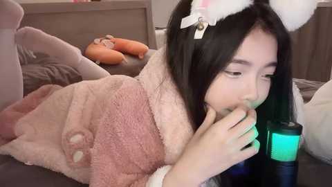 Media: A video of an Asian woman in a plush pink cat robe, wearing cat ears, sitting on a bed with orange pillows, holding a green drink, in a cozy indoor setting.