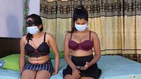 Media: Video of two young women with light brown skin, wearing face masks, black lace bras, and plaid skirts, sitting on a bed with green pillows, in a room with beige curtains.