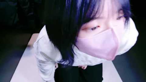 Media: A video of an Asian woman in a white shirt and black pants, wearing a pink mask, kneeling on a white mat, with a dark background, suggesting a dimly lit room.