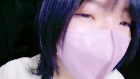 Media: Close-up video of an Asian woman with short, straight, dark blue hair, wearing a pink face mask, revealing only her eyes, nose, and mouth. Background is dark and indistinct.